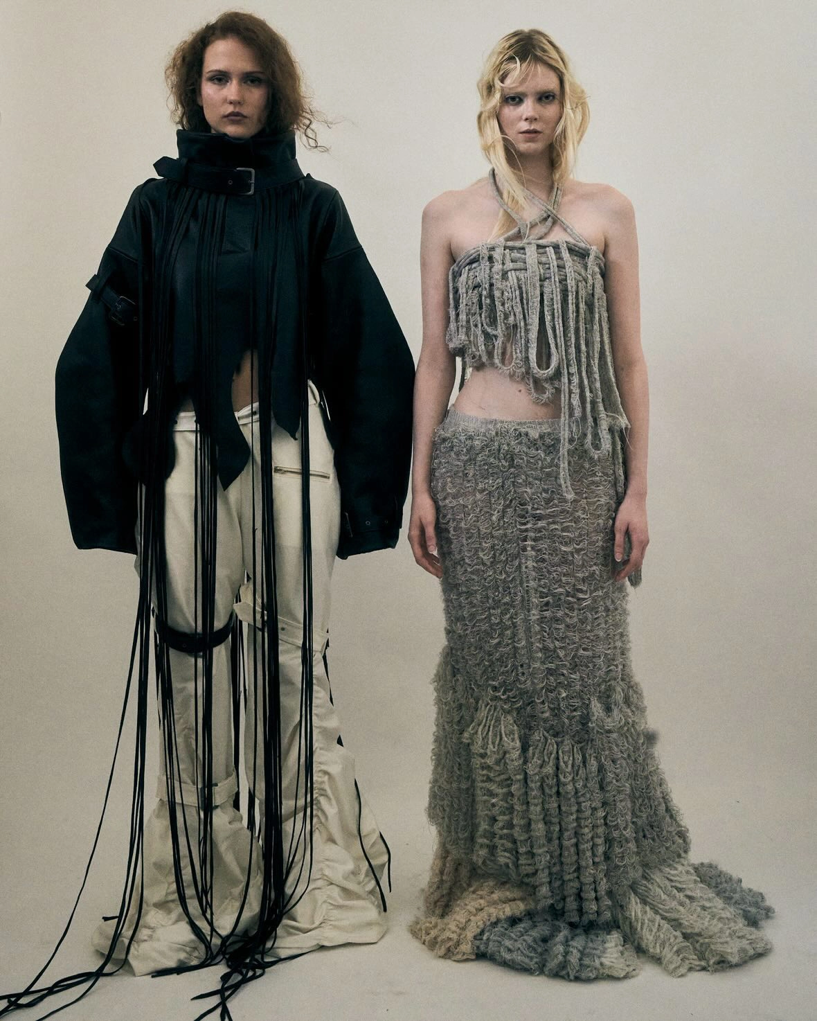 Two models wearing avant-garde graduate fashion designs featuring handwoven knitwear, cascading textile details, and oversized silhouettes against a neutral studio backdrop.